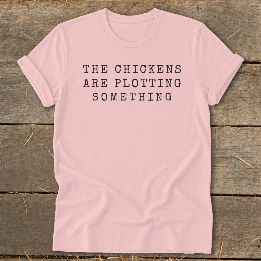 Pink t-shirt with black text reading "THE CHICKENS ARE PLOTTING SOMETHING".