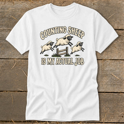 White t-shirt with cartoon sheep jumping over a fence and text "Counting Sheep is my actual job".