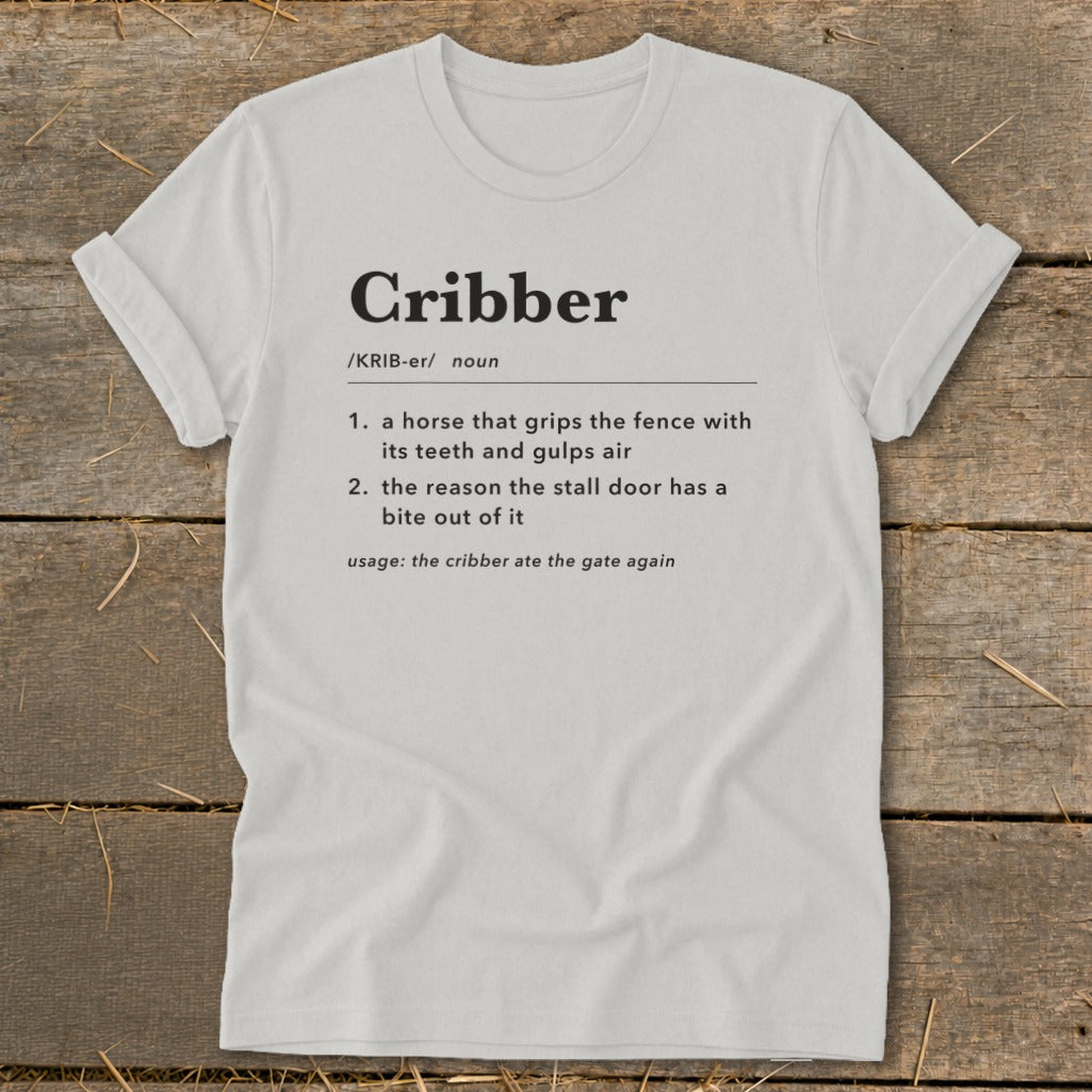 Light gray t-shirt featuring dictionary-style text defining the word Cribber on a wooden surface.