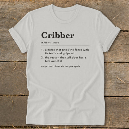 Light gray t-shirt featuring dictionary-style text defining the word Cribber on a wooden surface.