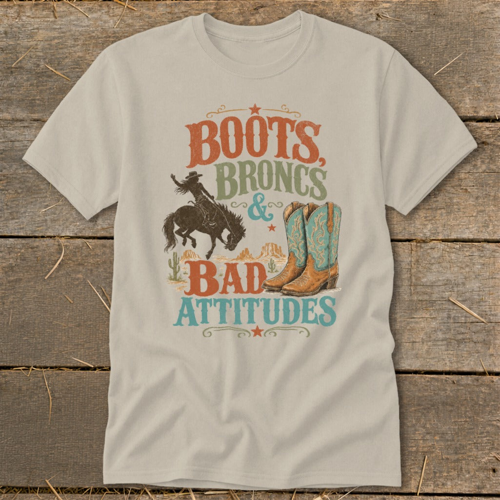 T-shirt graphic with text "Boots, Broncs & Bad Attitudes," cowboy, boots, and desert landscape.