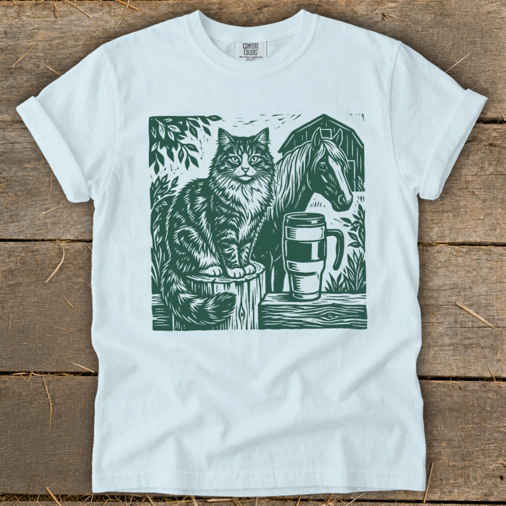 White t-shirt with green linocut print of a cat, horse, mug, and barn.