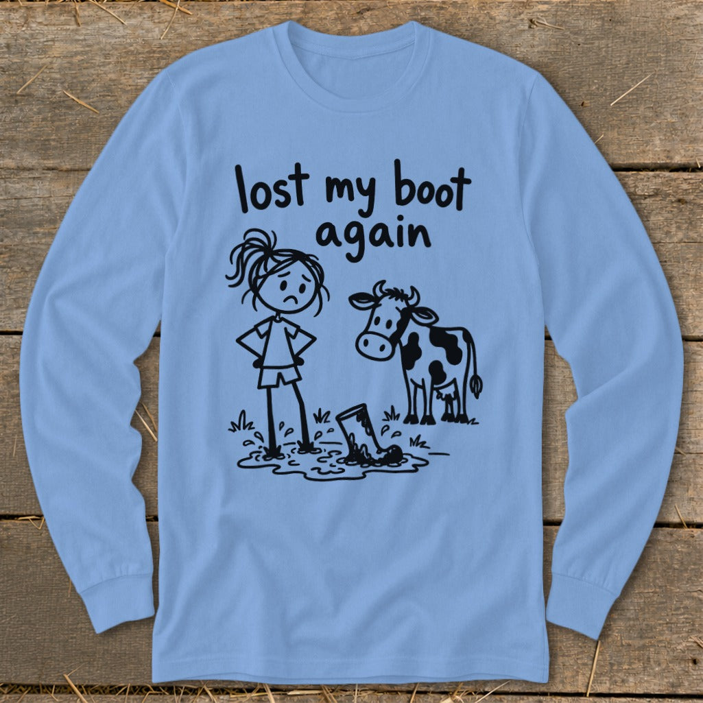 A light blue long-sleeve shirt featuring a black line drawing of a girl and a cow looking at a muddy boot, with the text "lost my boot again."