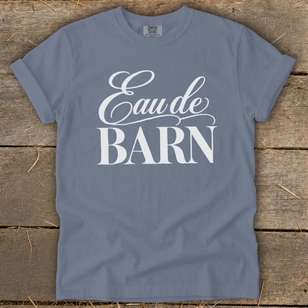 Blue t-shirt with white "Eau de BARN" text on a wooden background.