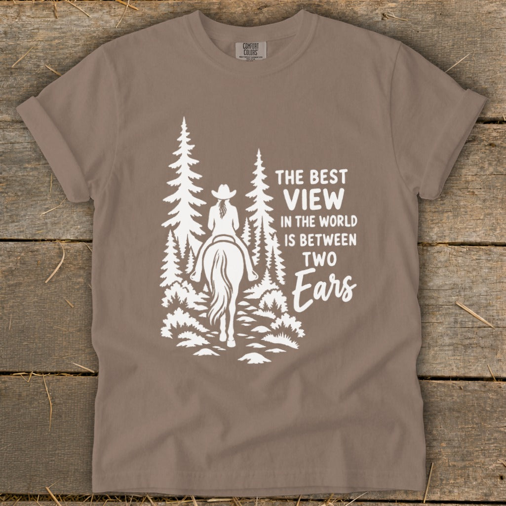 A taupe t-shirt featuring a white graphic of a horse rider in the forest with text that reads, "The best view in the world is between two ears."