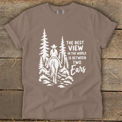 A taupe t-shirt featuring a white graphic of a horse rider in the forest with text that reads, "The best view in the world is between two ears."