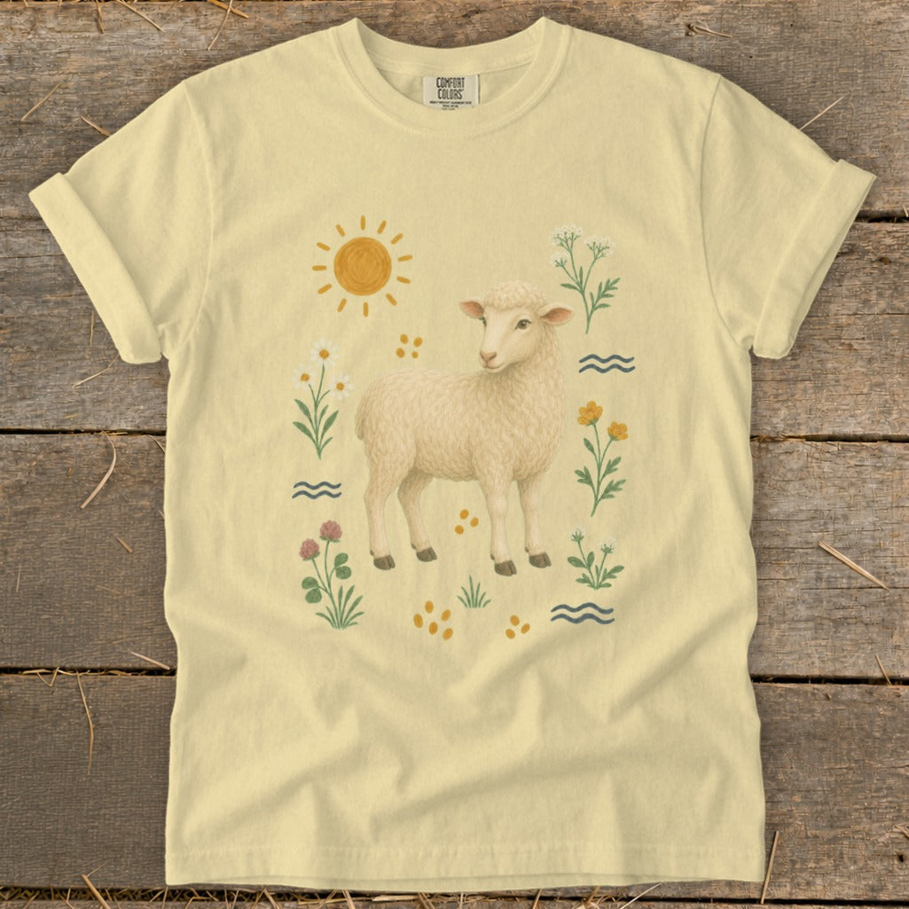 Cream t-shirt with a sheep, sun, flowers, and water on a wooden background.