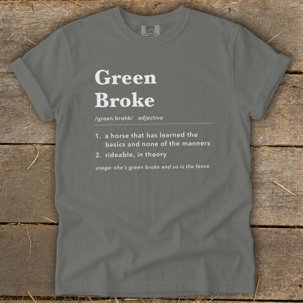 A dark gray t-shirt featuring white text defining the adjective "Green Broke" on a rustic wooden background.