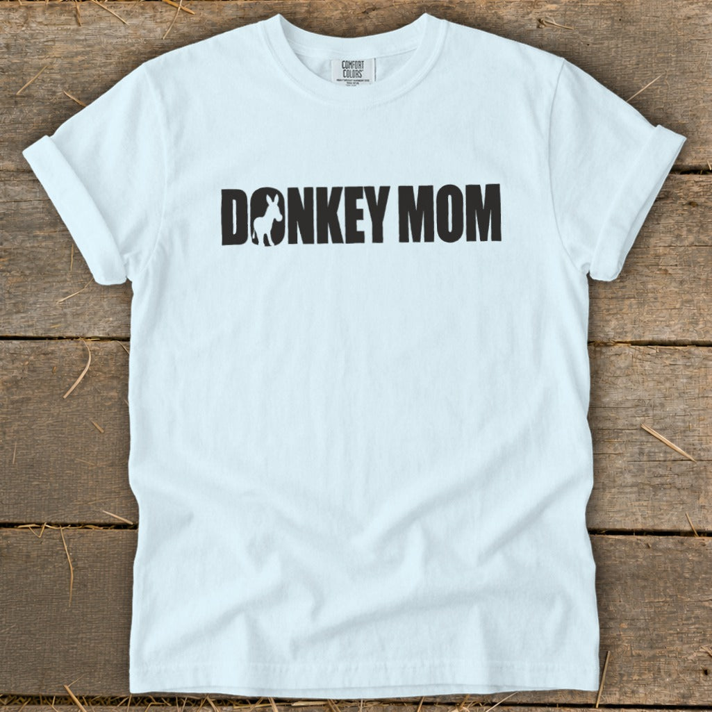 A light blue t-shirt with rolled sleeves lies flat on a wooden surface, featuring bold black text reading DONKEY MOM with a silhouette of a donkey inside the letter O.