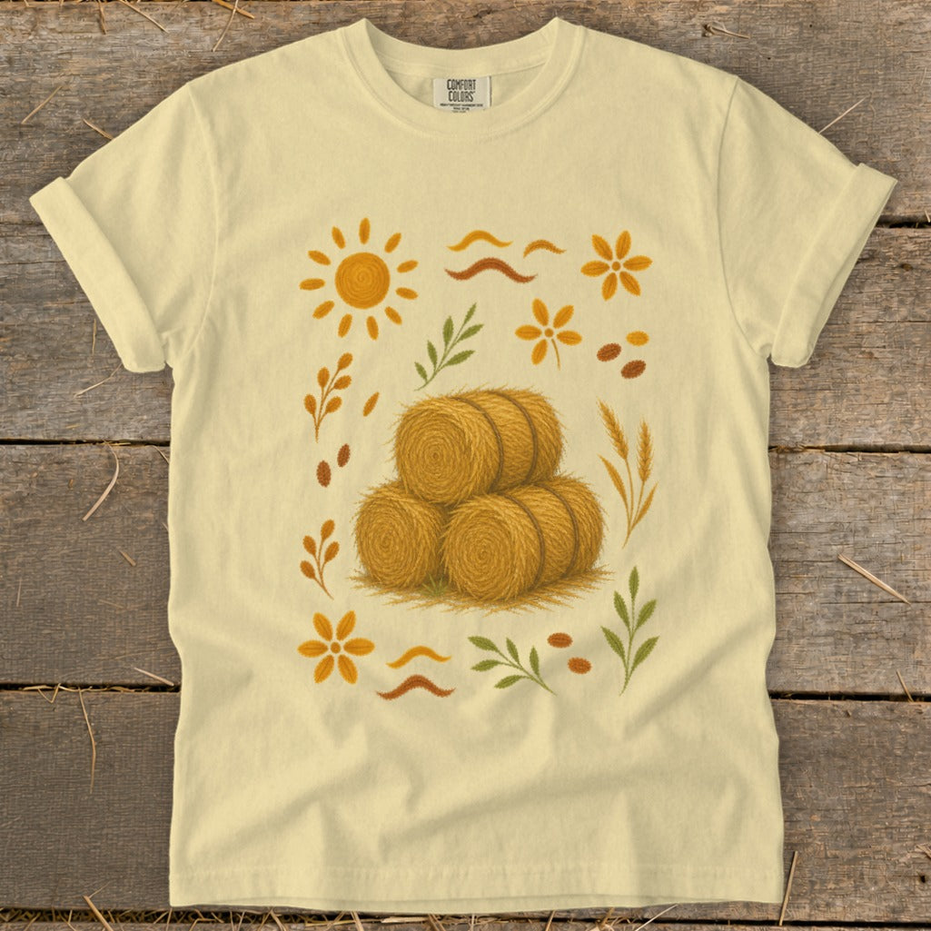 Light yellow t-shirt with a central illustration of three round hay bales surrounded by autumn motifs.
