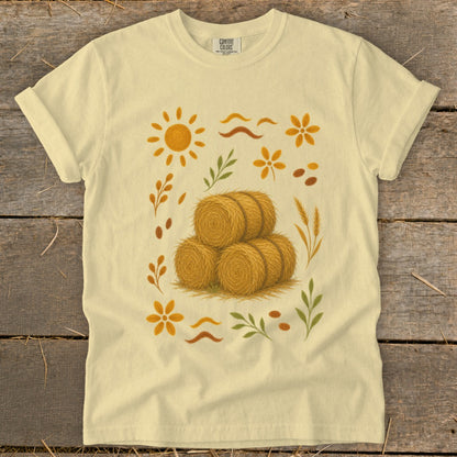 Light yellow t-shirt with a central illustration of three round hay bales surrounded by autumn motifs.
