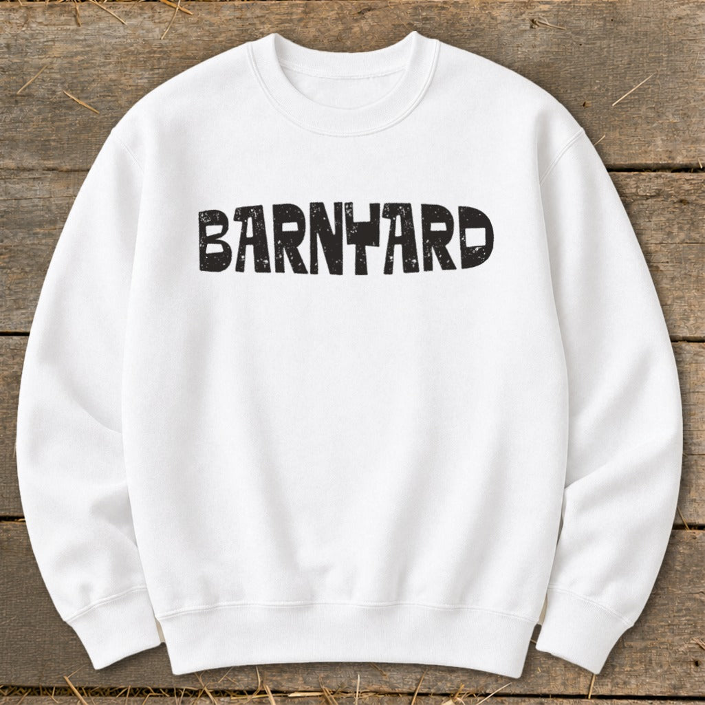 A white crewneck sweatshirt featuring the black distressed text BARNYARD centered across the chest, laid flat on a rustic wooden background.