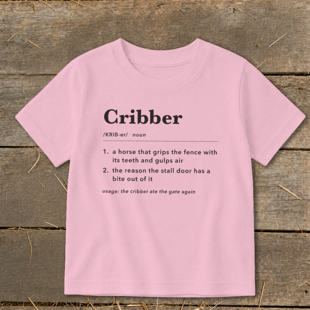 Light pink children's t-shirt printed with a humorous dictionary definition of the word cribber on a wooden surface.
