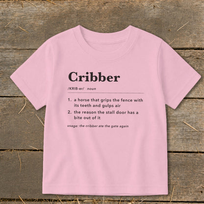 Light pink children's t-shirt printed with a humorous dictionary definition of the word cribber on a wooden surface.
