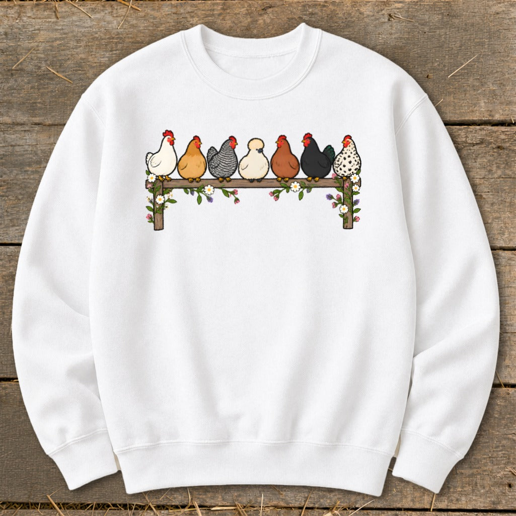 A white crewneck sweatshirt featuring a graphic of seven colorful cartoon chickens roosting on a wooden fence adorned with flowers.