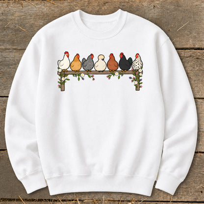 A white crewneck sweatshirt featuring a graphic of seven colorful cartoon chickens roosting on a wooden fence adorned with flowers.