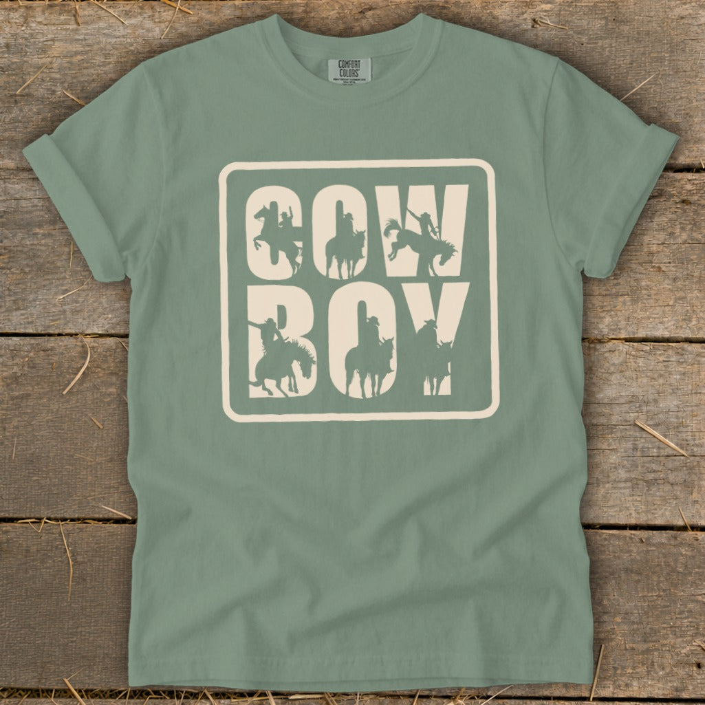Green t-shirt with a cream square graphic containing "COW" and "BOY" in large letters, filled with horse riders.