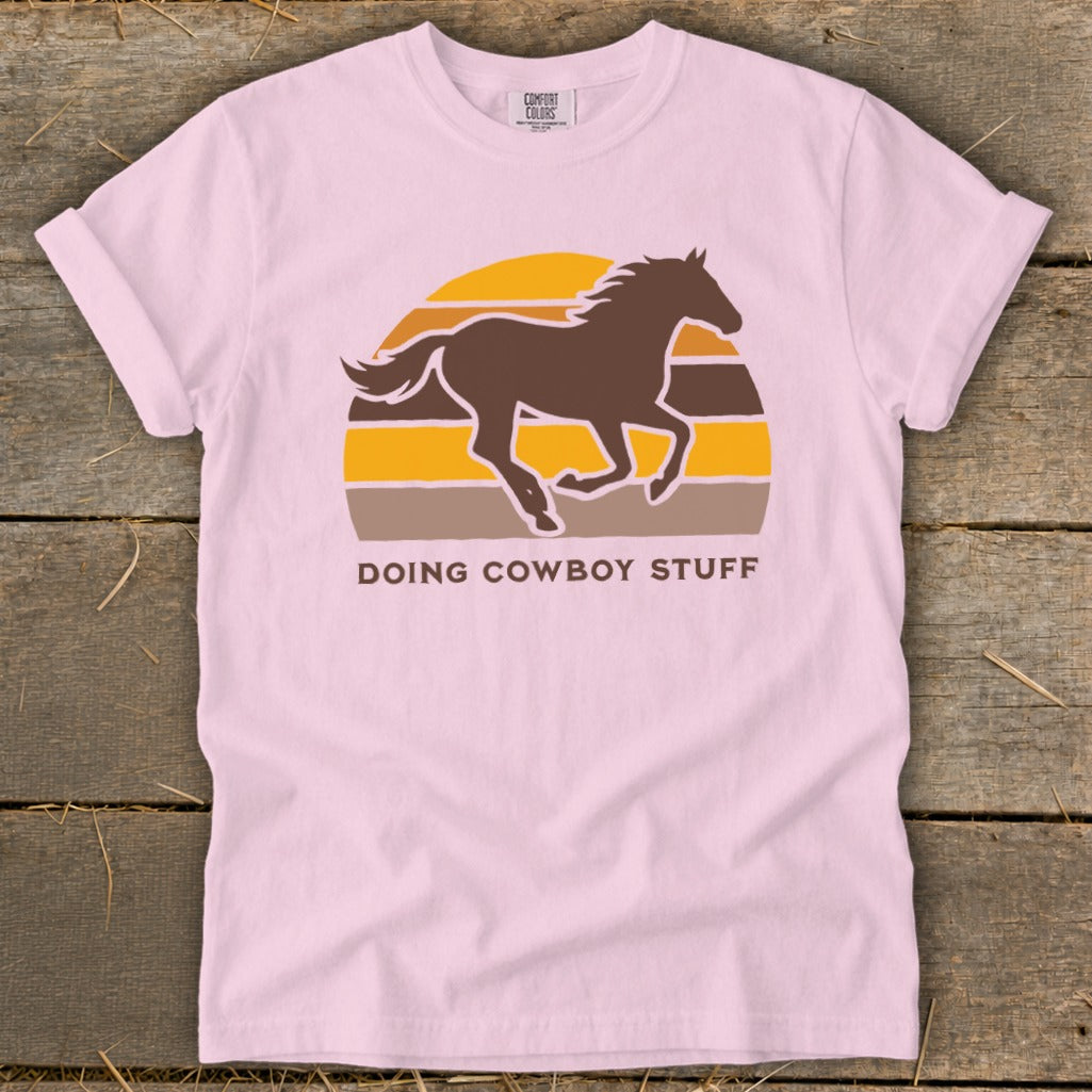 Pink t-shirt with a brown horse silhouette against a striped sunset and the text "DOING COWBOY STUFF".