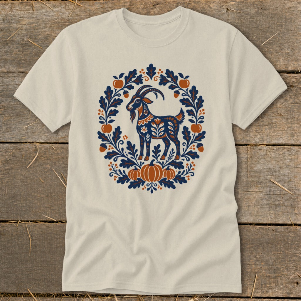 A cream-colored t-shirt with a blue and orange folk art illustration of a goat framed by leaves, acorns, and pumpkins.