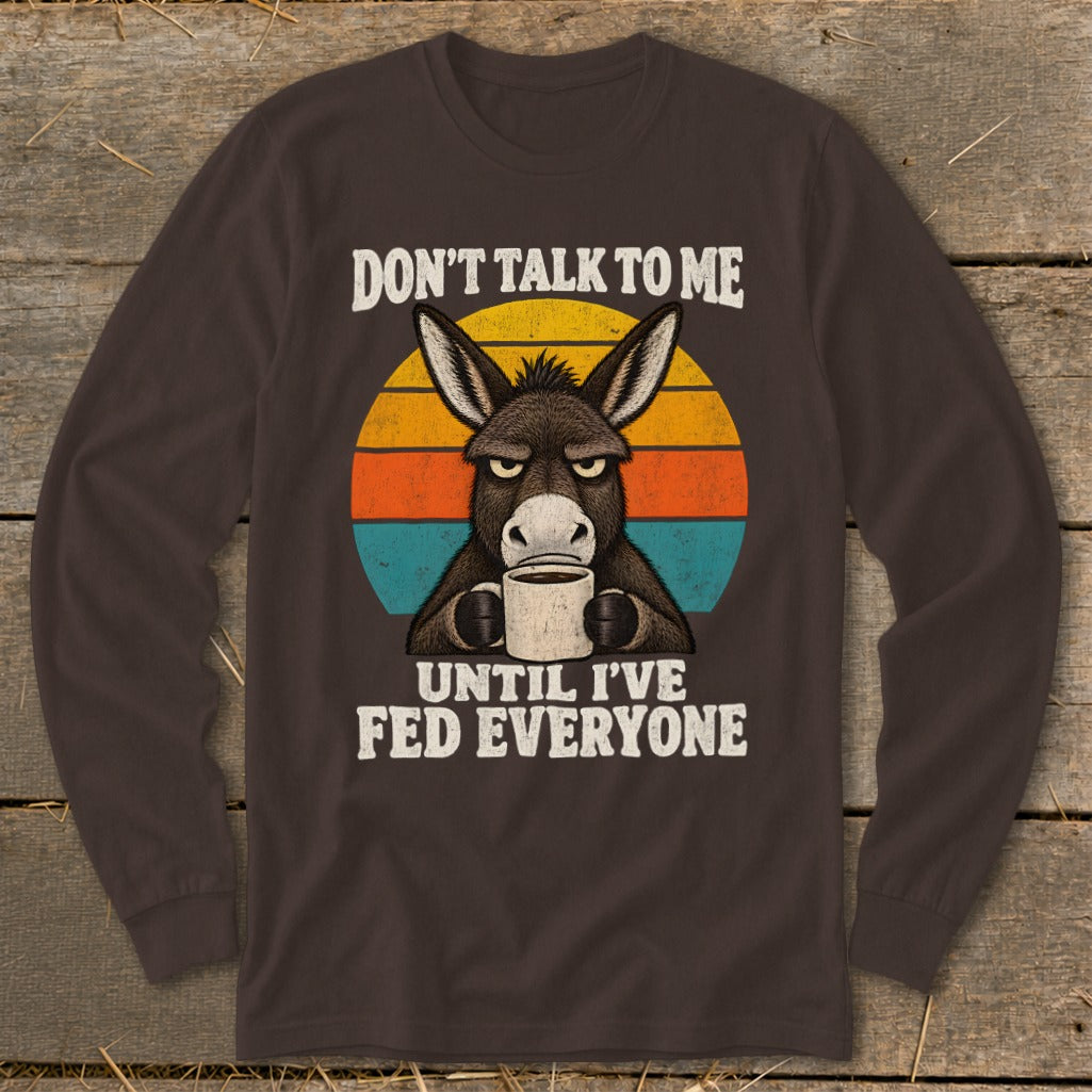 A dark brown long-sleeve t-shirt featuring an illustration of an annoyed donkey holding a coffee mug against a retro sunset graphic with the text DON'T TALK TO ME UNTIL I'VE FED EVERYONE.