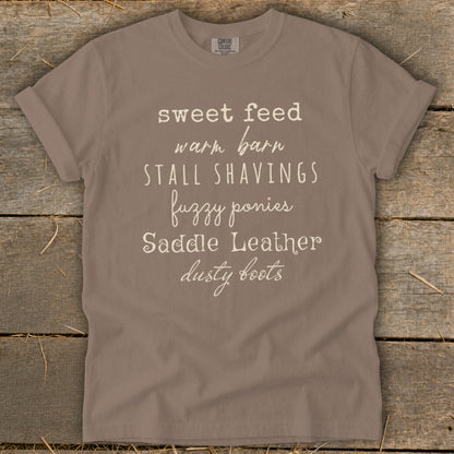 Taupe t-shirt with white text: sweet feed, warm barn, stall shavings, fuzzy ponies, saddle leather, dusty boots.