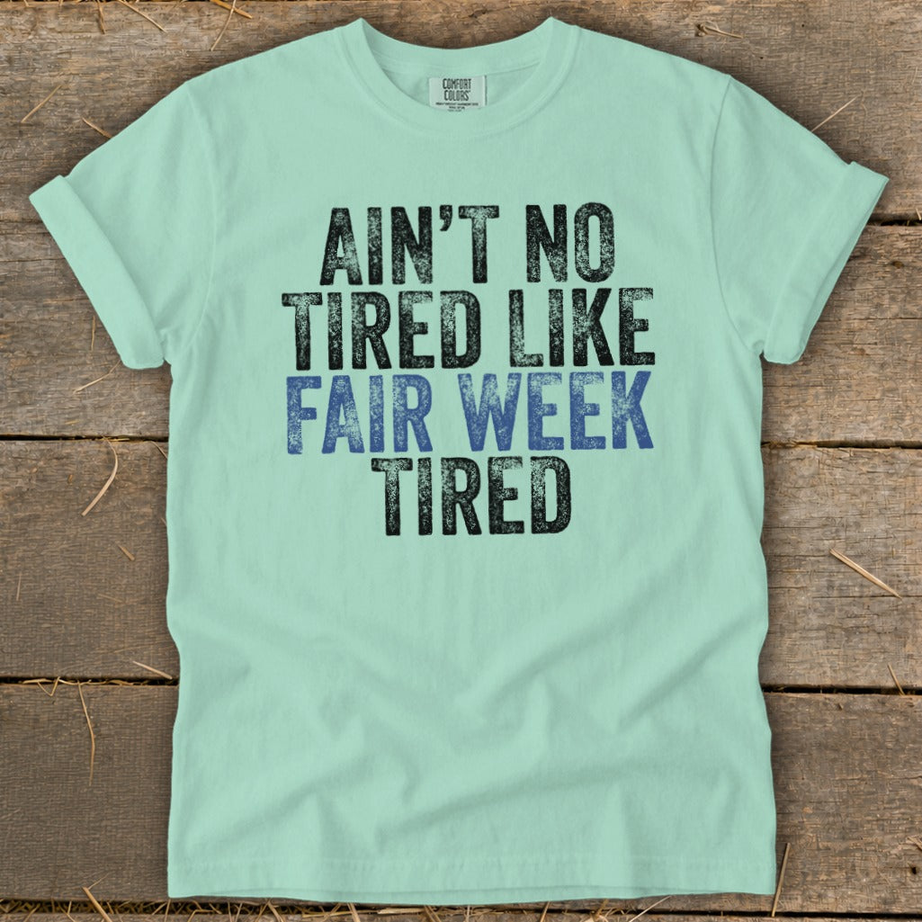Light blue t-shirt with black and blue text reading "Ain't no tired like fair week tired".