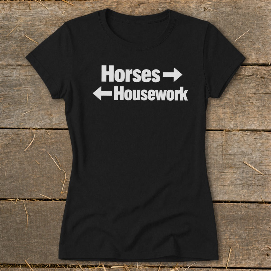 A black women's t-shirt rests on a wooden surface, featuring the white text Horses with a right arrow above Housework with a left arrow.