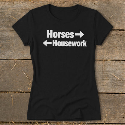 A black women's t-shirt rests on a wooden surface, featuring the white text Horses with a right arrow above Housework with a left arrow.