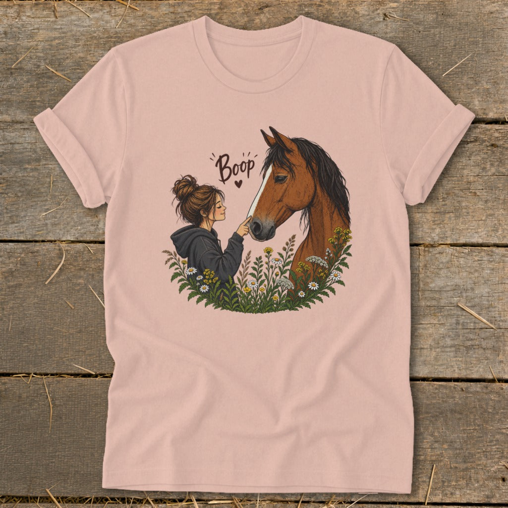 A pink t-shirt featuring an illustration of a woman touching a horse's nose amid wildflowers with the text Boop.