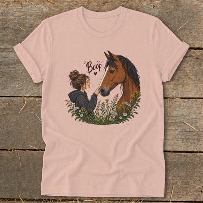 A pink t-shirt featuring an illustration of a woman touching a horse's nose amid wildflowers with the text Boop.