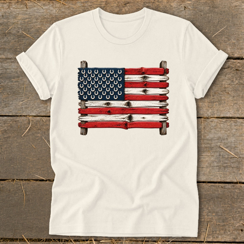 A cream-colored t-shirt on a wooden background, featuring an American flag made of painted wooden planks and horseshoes.