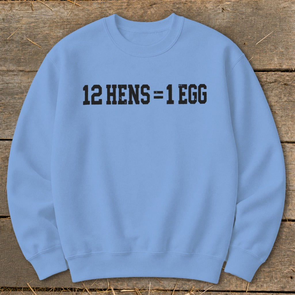 Light blue crewneck sweatshirt featuring the black distressed text 12 HENS = 1 EGG laid flat on a rustic wooden background.