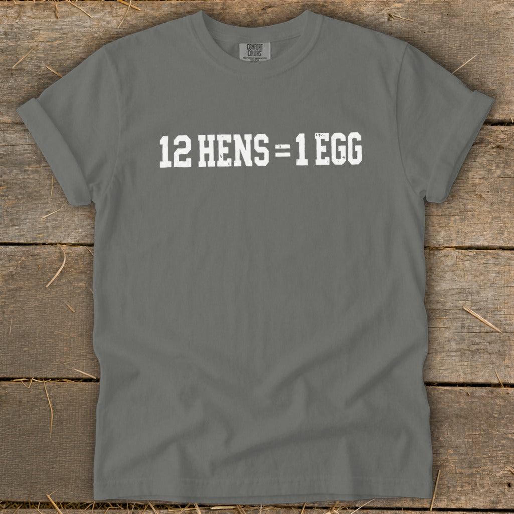 A grey short-sleeve t-shirt with distressed white text reading 12 HENS = 1 EGG, displayed on a wooden surface.