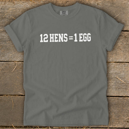 A grey short-sleeve t-shirt with distressed white text reading 12 HENS = 1 EGG, displayed on a wooden surface.