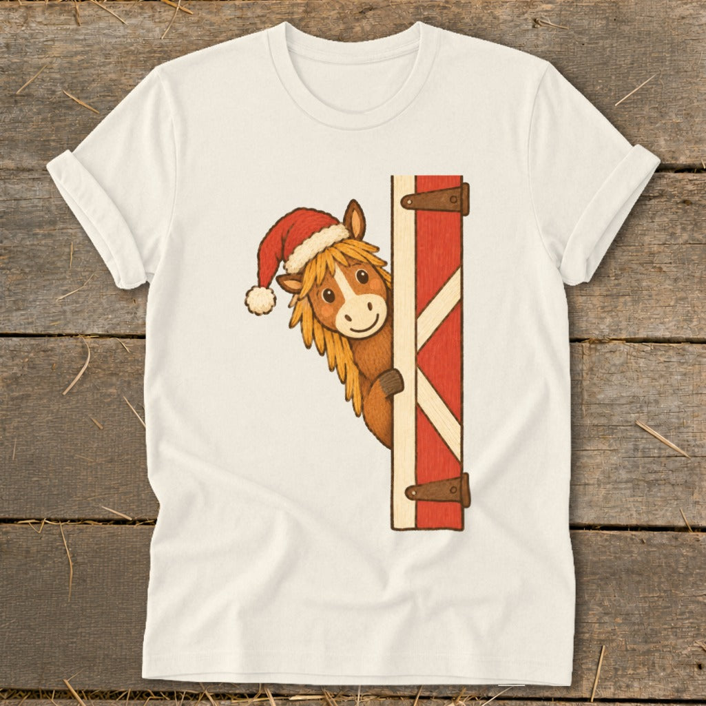 Horse wearing a Santa hat peeking around a red barn door on a cream t-shirt.
