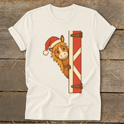 Horse wearing a Santa hat peeking around a red barn door on a cream t-shirt.