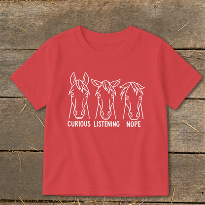 A red children's t-shirt displays white line drawings of three horse heads labeled Curious, Listening, and Nope.