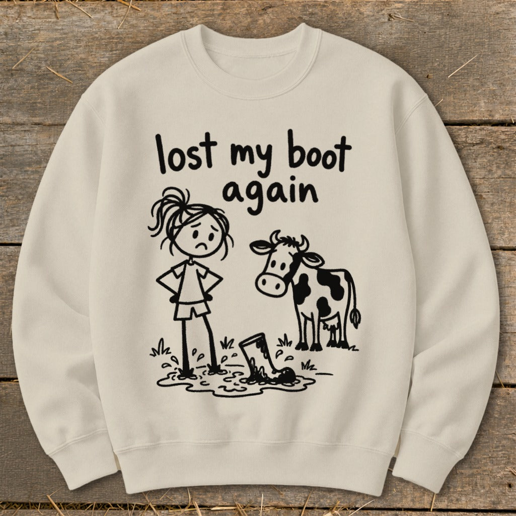 A cream-colored crewneck sweatshirt features a black cartoon illustration of a distressed girl and a cow looking at a muddy boot, with the text "lost my boot again."