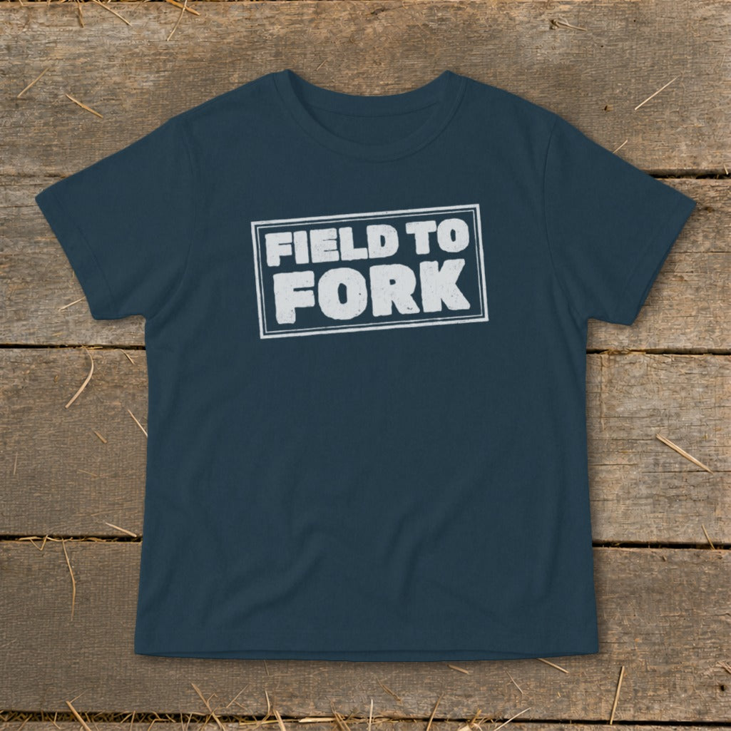 Dark blue short-sleeve t-shirt laid flat on a rustic wooden surface, featuring the white text "FIELD TO FORK" enclosed in a distressed rectangular outline on the chest.