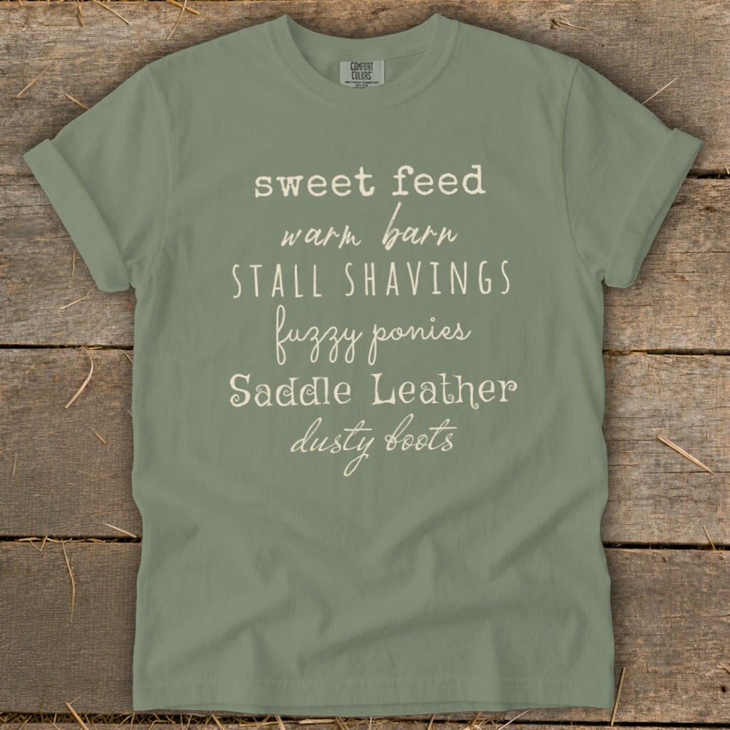 Sage t-shirt with white text: sweet feed, warm barn, stall shavings, fuzzy ponies, saddle leather, dusty boots.