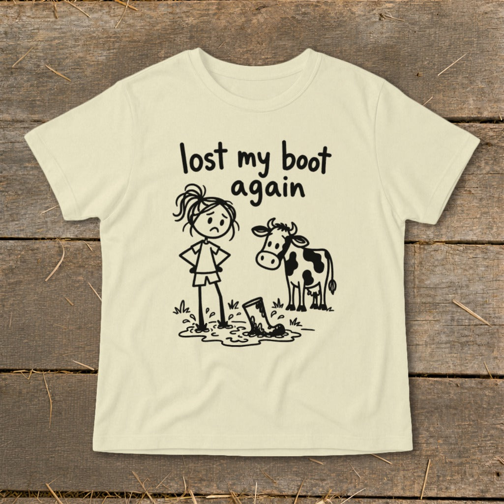 A cream-colored t-shirt with a graphic of a sad girl and a cow looking at a boot stuck in mud, accompanied by the text lost my boot again.