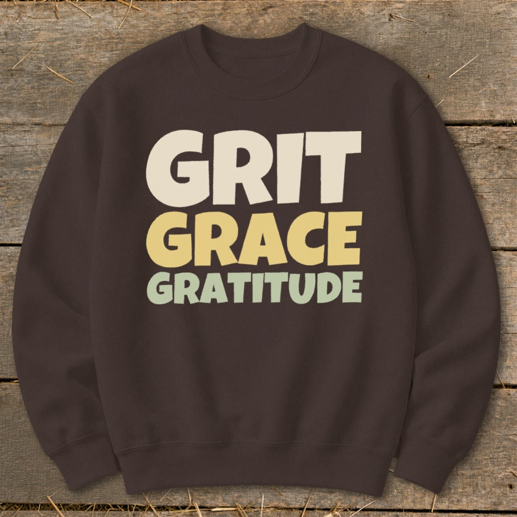 A dark brown crewneck sweatshirt featuring the text GRIT GRACE GRATITUDE printed centrally in cream, yellow, and green block letters.
