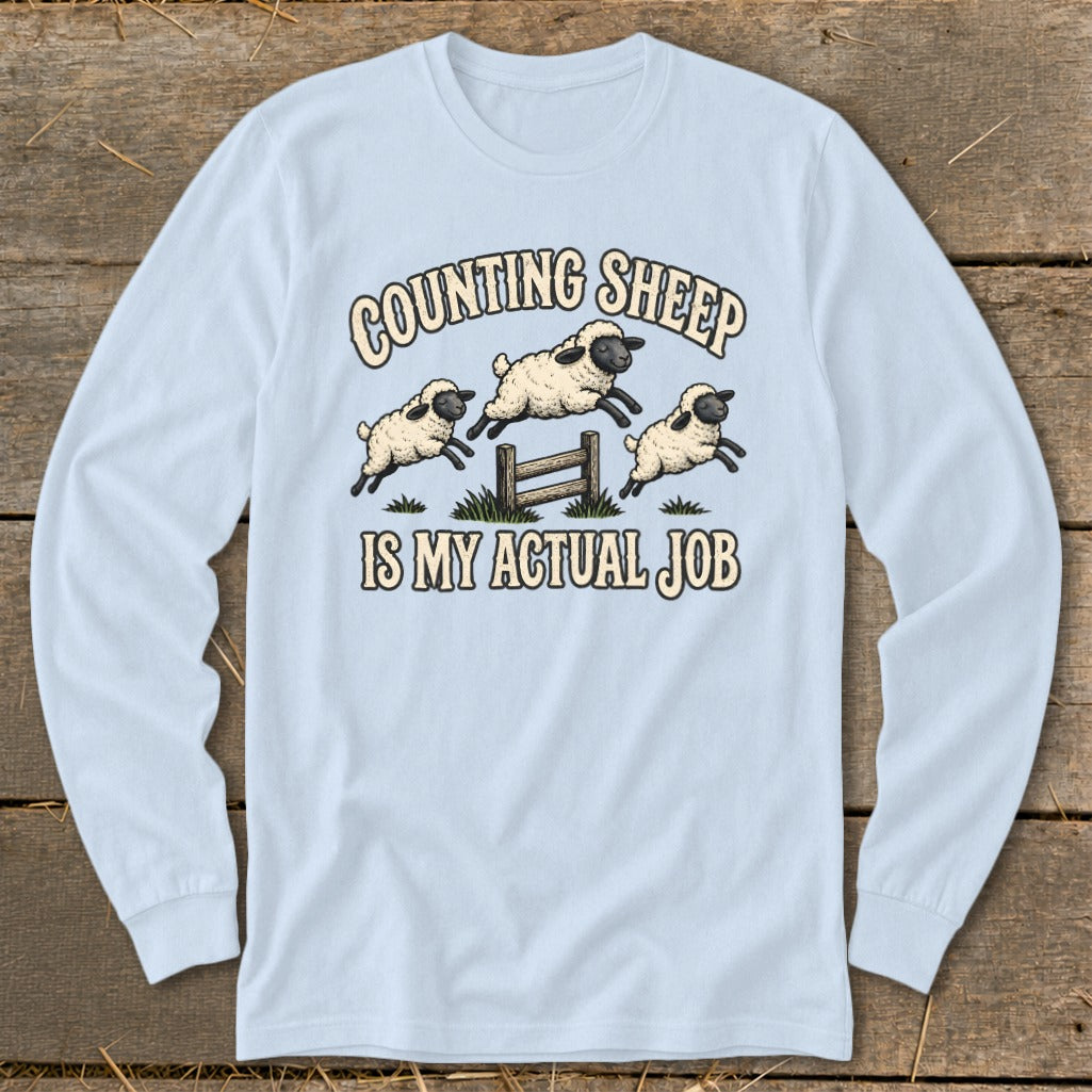 Light blue long-sleeve shirt with cartoon sheep jumping over a fence with text "Counting Sheep Is My Actual Job".