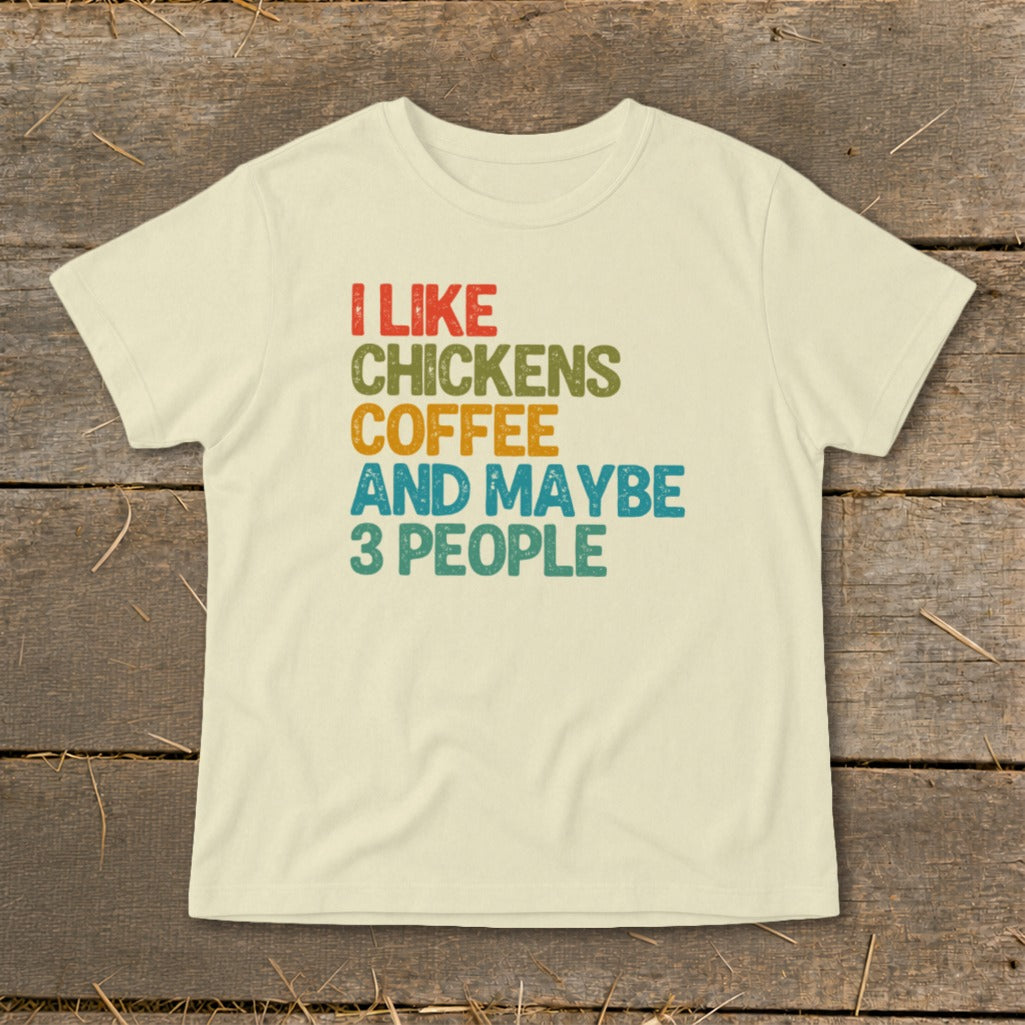 A cream-colored short-sleeved t-shirt with distressed, colorful text reading I LIKE CHICKENS COFFEE AND MAYBE 3 PEOPLE centered on the front, displayed flat on a rustic wooden surface.