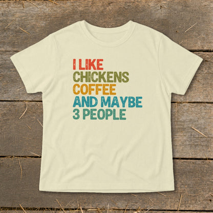 A cream-colored short-sleeved t-shirt with distressed, colorful text reading I LIKE CHICKENS COFFEE AND MAYBE 3 PEOPLE centered on the front, displayed flat on a rustic wooden surface.