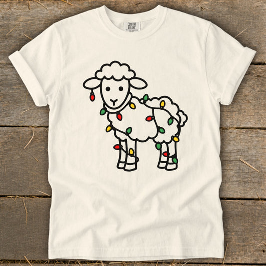 A cream-colored t-shirt on a wooden surface displays a black line drawing of a sheep wrapped in colorful holiday lights.