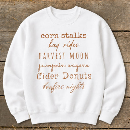 White sweatshirt with text listing fall activities: corn stalks, hay rides, harvest moon, pumpkin wagons, cider donuts, bonfire nights.