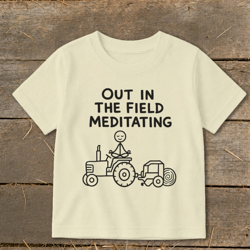 Light yellow t-shirt with black text "OUT IN THE FIELD MEDITATING" and tractor graphic.