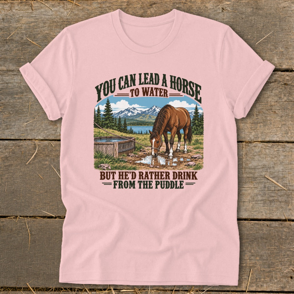 Pink t-shirt with a vintage graphic of a horse drinking from a puddle, featuring mountains and the text "You can lead a horse to water, but he'd rather drink from the puddle."