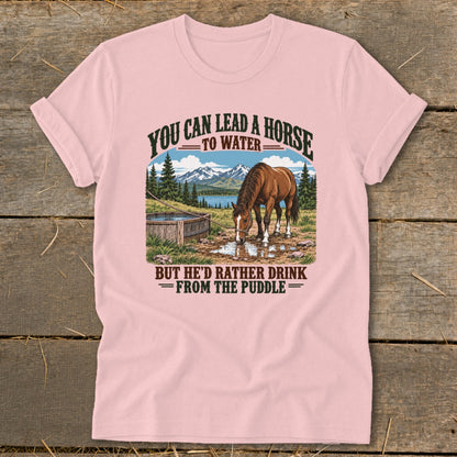 Pink t-shirt with a vintage graphic of a horse drinking from a puddle, featuring mountains and the text "You can lead a horse to water, but he'd rather drink from the puddle."